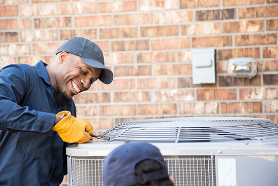 Technician smiling while servicing outdoor AC unit next to brick home.