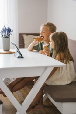 Two young kids sitting at a table eating ice cream cones, watching something on a tablet.